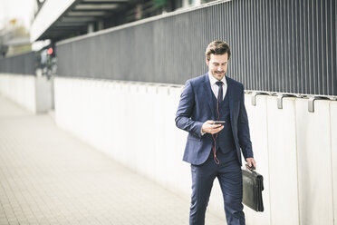 Businessman walking in the city with cell phone and earphones - UUF17679