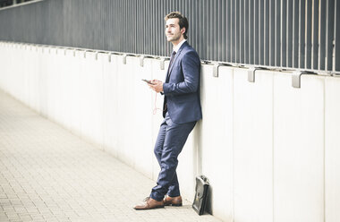 Businessman leaning against a wall in the city with cell phone and earphones - UUF17681