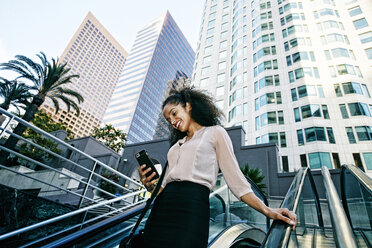 Smiling Hispanic businesswoman texting on escalator outdoors - BLEF04230