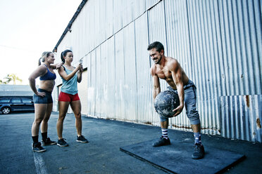 Women watching man lifting heavy ball outdoors - BLEF04272