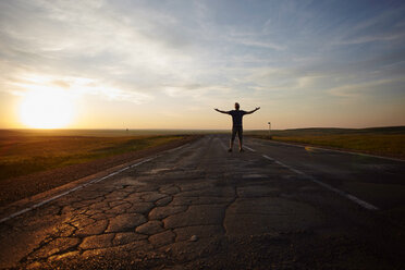 Caucasian man standing in cracked road at sunset with arms outstretched - BLEF04328