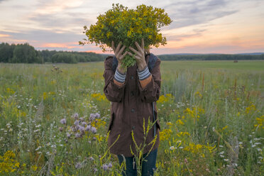 Kaukasische Frau hält Blumenstrauß über Gesicht in Feld bei Sonnenuntergang - BLEF04343