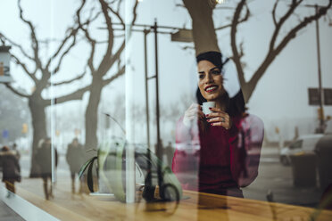 Smiling young woman with cup of coffee behind windowpane in a cafe - ACPF00510