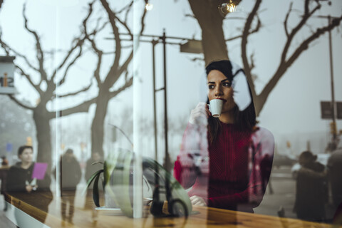 Junge Frau trinkt eine Tasse Kaffee hinter einer Fensterscheibe in einem Café, lizenzfreies Stockfoto