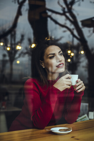 Lächelnde junge Frau mit einer Tasse Kaffee hinter einer Fensterscheibe in einem Cafe, lizenzfreies Stockfoto