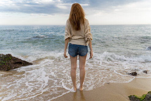 Rückenansicht einer rothaarigen jungen Frau, die am Strand steht und auf das Meer schaut - AFVF02984