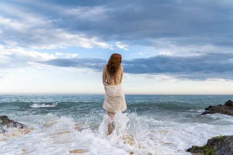 Rückenansicht einer rothaarigen jungen Frau ohne Hemd, die vor dem Meer steht, lizenzfreies Stockfoto