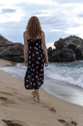 Back view of young woman wearing summer dress with floral design walkung on the beach - AFVF03016