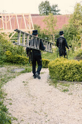 Two chimney sweeps walking with ladder on garden path - MAEF12886