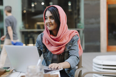 Lächelnde junge Frau mit Laptop und Kopftuch in einem Straßencafé - OCAF00396