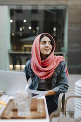 Smiling young woman with laptop wearing headscarf at a pavement cafe - OCAF00397