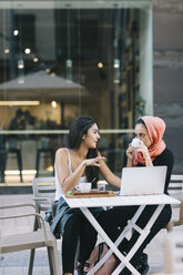 Zwei Freunde sitzen in einem Straßencafé mit Laptop zusammen und unterhalten sich - OCAF00400