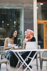 Two happy friends sitting together at a pavement cafe with laptop - OCAF00403