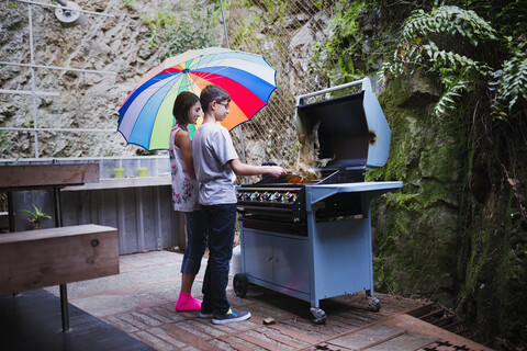 Mixed Race Bruder und Schwester kochen auf der Terrasse Grill unter Sonnenschirm, lizenzfreies Stockfoto