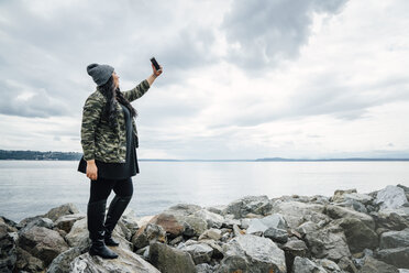 Gemischtrassige Frau steht auf Felsen am Meer und posiert für ein Selfie mit dem Handy - BLEF04580
