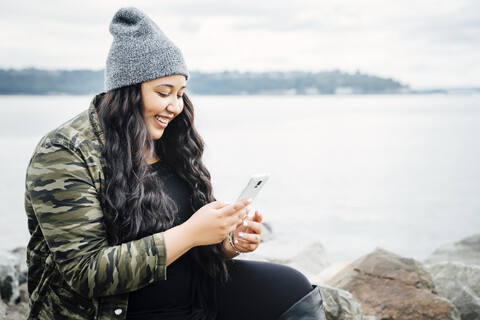 Lächelnde Mixed Race Frau texting auf Handy in der Nähe von Meer, lizenzfreies Stockfoto