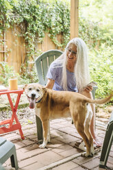 Caucasian woman holding cell phone petting dog on backyard patio - BLEF04902