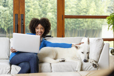 Lächelnde schwarze Frau streichelt weißen Hund auf Sofa mit Laptop - BLEF05114