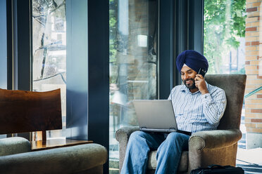 Mann mit Turban, der in einem Sessel Laptop und Mobiltelefon benutzt - BLEF05309