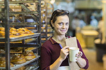 Kaukasische Frau hält Klemmbrett und trinkt Kaffee in einer Bäckerei - BLEF05547