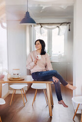 Mature woman sitting on kitchen table, drinking coffee - HAPF02893