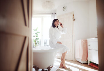 Mature woman sitting on bath tub, applying mascara - HAPF02940