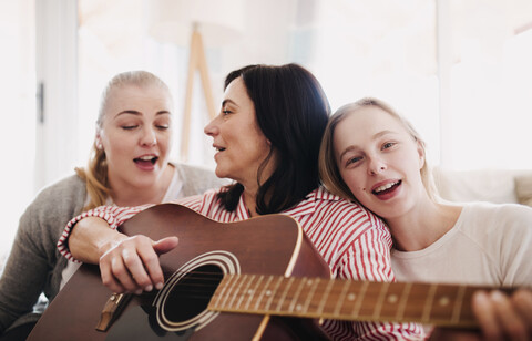 Reife Frau mit zwei Töchtern singt und spielt Gitarre zu Hause, lizenzfreies Stockfoto