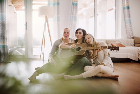 Mature woman with two daughters sitting on the floor playing guitar at home - HAPF02965