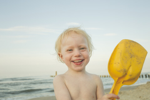 Porträt eines glücklichen kleinen Mädchens, das mit einer gelben Schaufel am Strand spielt, lizenzfreies Stockfoto