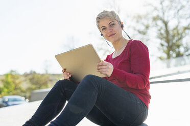 Portrait of young freelancer sitting outdoors using digital tablet and headset - FBAF00599