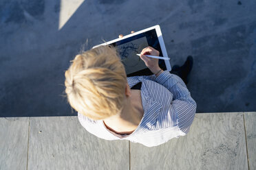 Young woman leaning against wall drawing on digital tablet, top view - FBAF00603