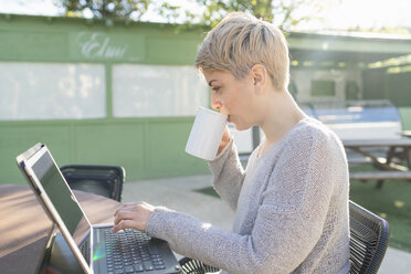 Junge Frau trinkt eine Tasse Kaffee im Freien, während sie einen Laptop benutzt - FBAF00610