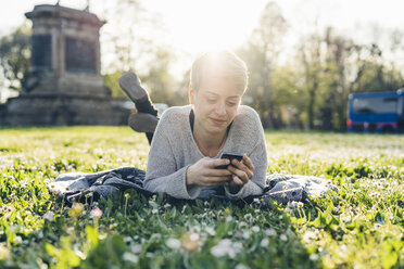 Smiling young woman lying on a meadow at backlight looking at cell phone - FBAF00612