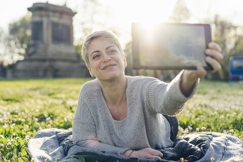 Porträt einer glücklichen jungen Frau, die auf einer Wiese liegt und ein Selfie mit einem digitalen Tablet macht, lizenzfreies Stockfoto