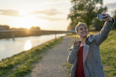 Porträt einer grinsenden Frau, die bei Sonnenuntergang ein Selfie mit ihrem Smartphone macht - FBAF00627