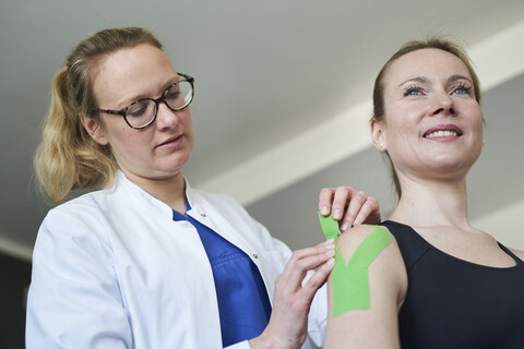 Ärztin beim Anlegen eines Pflasters an der Schulter eines Patienten in einer Arztpraxis, lizenzfreies Stockfoto