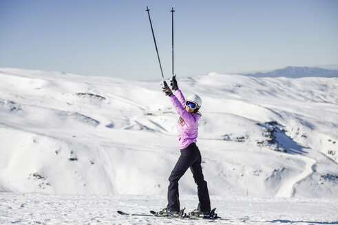 Glückliche Frau, die ihre Skistöcke in einer schneebedeckten Landschaft in der Sierra Nevada, Andalusien, Spanien, hochhält - JSMF01101
