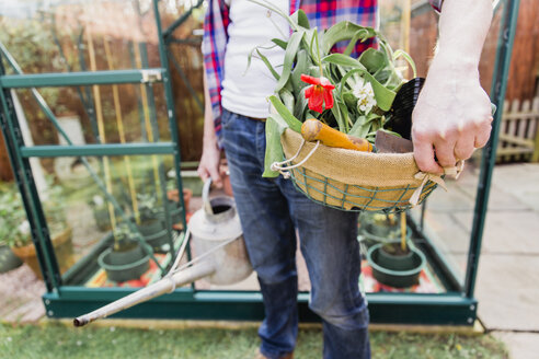 Close-up of man holding basket with flowers and watering can before greenhouse in garden - NMS00320