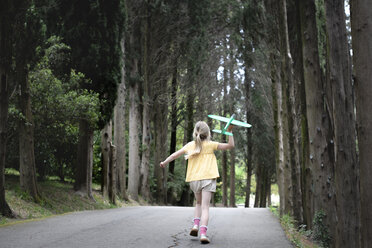 Mädchen mit Spielzeugflugzeug läuft auf einer mit Bäumen gesäumten Straße - EYAF00250