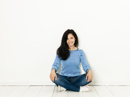 Schöne junge Frau mit schwarzen Haaren und blau-weiß gestreiften Pullover sitzt auf dem Boden vor weißem Hintergrund - HMEF00415