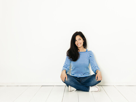 Beautiful young woman with black hair and blue white striped sweater sitting on the ground in front of white background stock photo