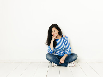 Schöne junge Frau mit schwarzen Haaren und blau-weiß gestreiften Pullover sitzt auf dem Boden vor weißem Hintergrund - HMEF00417