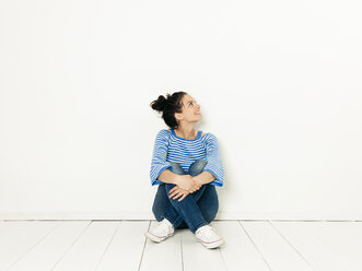 Beautiful young woman with black hair and blue white striped sweater sitting on the ground in front of white background - HMEF00418