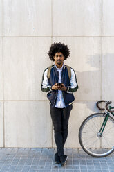 Portrait of casual businessman with bicycle and cell phone standing at a wall - AFVF03131