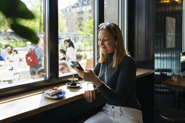 Lächelnde junge Frau in einem Café, die auf ihr Smartphone schaut - FBAF00634