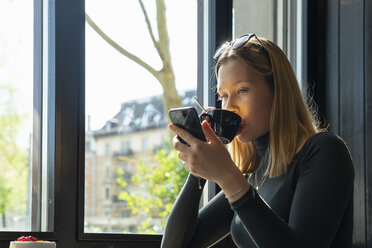 Young woman in a coffee shop drinking coffee while looking at smartphone - FBAF00635