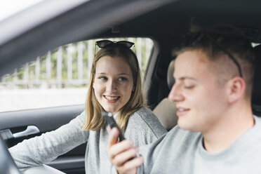 Portrait of young woman in a car watching her boyfriend on driver's seat using smartphone - FBAF00640