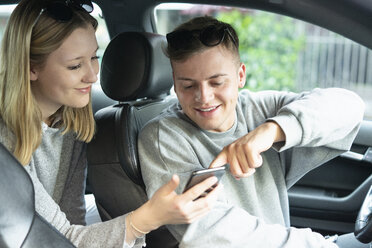 Lächelndes junges Paar in einem Auto, das auf ein Mobiltelefon schaut - FBAF00644