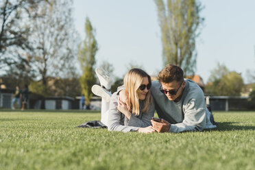 Junges Paar mit Sonnenbrille liegt auf einer Wiese und schaut auf sein Handy - FBAF00658