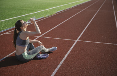 Sportswoman sitting on racetrack and drinking water after workout - AHSF00466